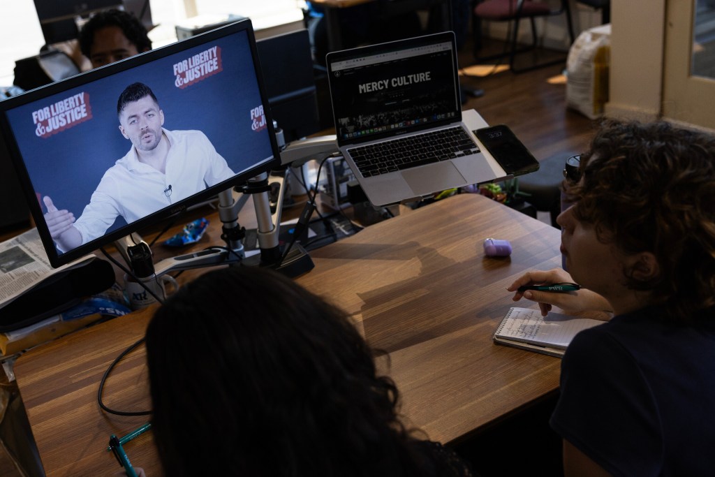 Two people seen from behind look at a monitor, which shows a man in a white shirt speaking in front of a backdrop that reads “For Liberty & Justice.”