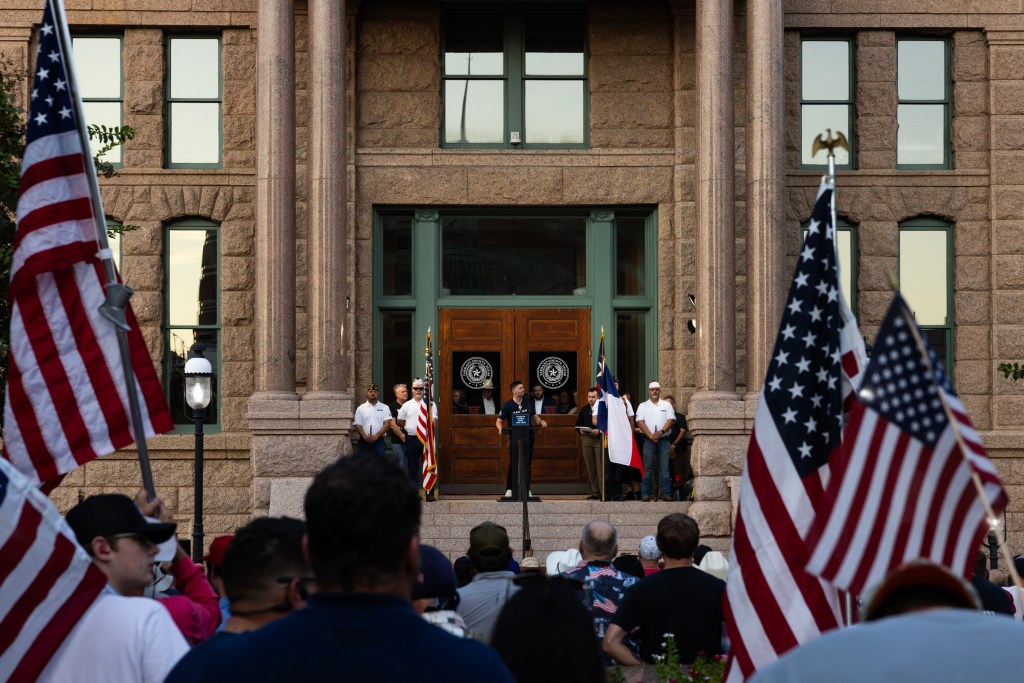 A man, seen from the back of a crowd, stands on the steps of a stone building behind a small lectern. Multiple people in the crowd are waving large American flags.